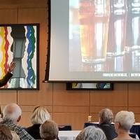 A man gestures to a screen with 4 glasses of beer on it, while 20 people are seated at tables listening
